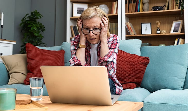 An older woman holding her head in shock as she looks at a laptop.