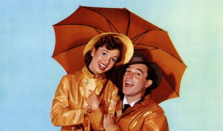 Lobby card artwork for “Singin’ in the Rain” (1952) showing Debbie Reynolds and Gene Kelly smiling under a large orange umbrella against a pale blue background. Both wear shiny yellow raincoats; she has a yellow rain hat and he wears a brown fedora.