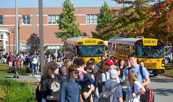 High school students walk along a sidewalk outside a brick school building as two yellow school buses wait under trees with fall foliage.
