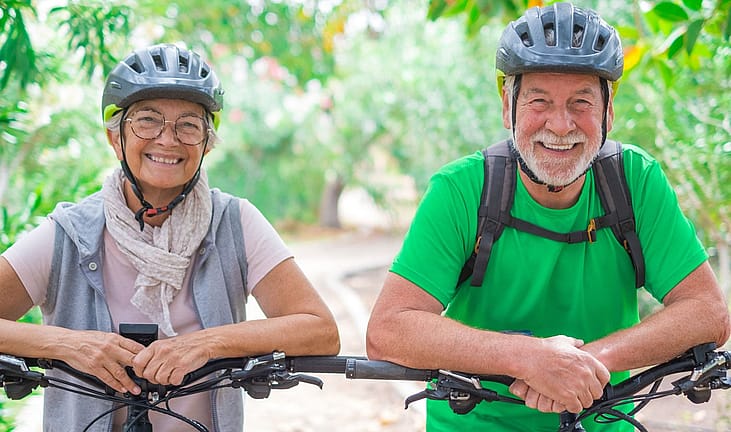 Smiling older couple in bike helmets leaning on their handlebars on a tree-lined path