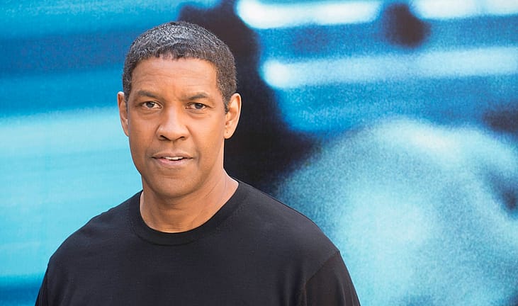 Denzel Washington in a black t-shirt in front of a blue background.
