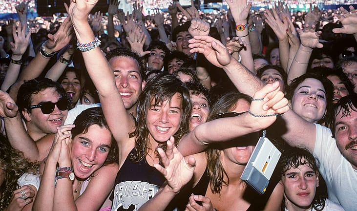 A crowd of smiling young fans with raised hands and friendship bracelets presses toward the stage at a stadium concert.