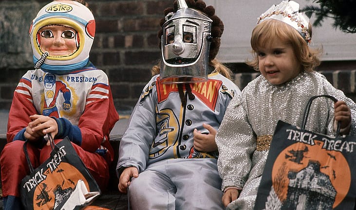 Three children in 1970 Halloween costumes sit on a brick stoop holding Trick or Treat bags: one in an astronaut mask and red smock, one in a silver Tin Man mask, and a smiling girl in a sequined princess dress and crown.