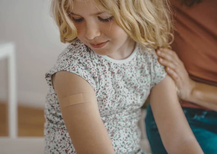 A young child with a small adhesive bandage on their upper arm. The child is wearing a floral-patterned shirt and looking down at the bandage.