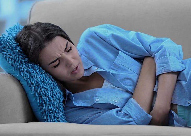 Young woman lying on sofa with closed eyes and holding her stomach.