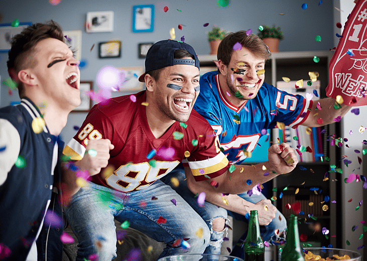 Three young men wearing football jerseys cheering while watching game at home with colorful confetti falling in the foreground. 