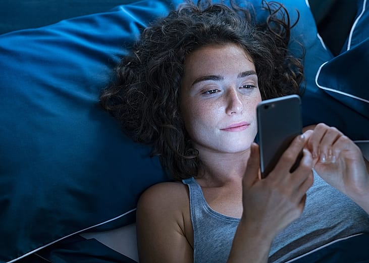 Young woman lies in bed in a dark room while looking at her phone.