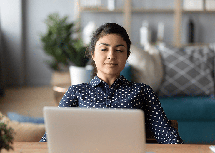 A calm-looking woman sitting with her eyes gently closed in front of a laptop in a home office.