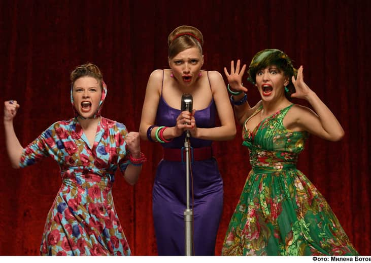 Three women wearing 50's era dresses onstage with a microphone.