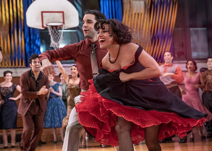 A woman in a black and red dress dancing and smiling with a man in khakis and a maroon shirt in a gym full of people.
