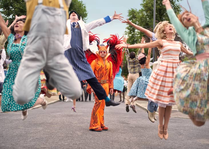 People singing and jumping up in the air next to Elton John's character in a red fire suit with horns and wings.