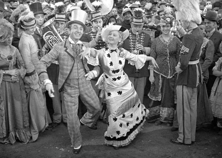 A man and woman singing with a band and crowd behind them.