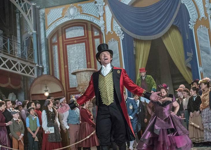 Hugh Jackman in a red coat and tails singing in a theater surrounded by people.