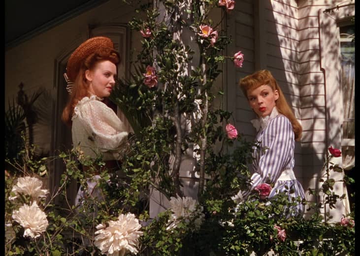 Judy Garland and another woman on the porch of a house peeking around a blooming vine.