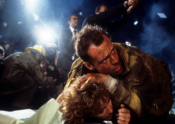 A man covered in black soot stands over a woman to protect her while fireman run around in background.