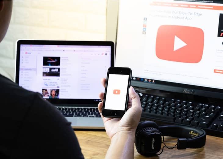 Person watching YouTube on a phone with two computer screens in the background.