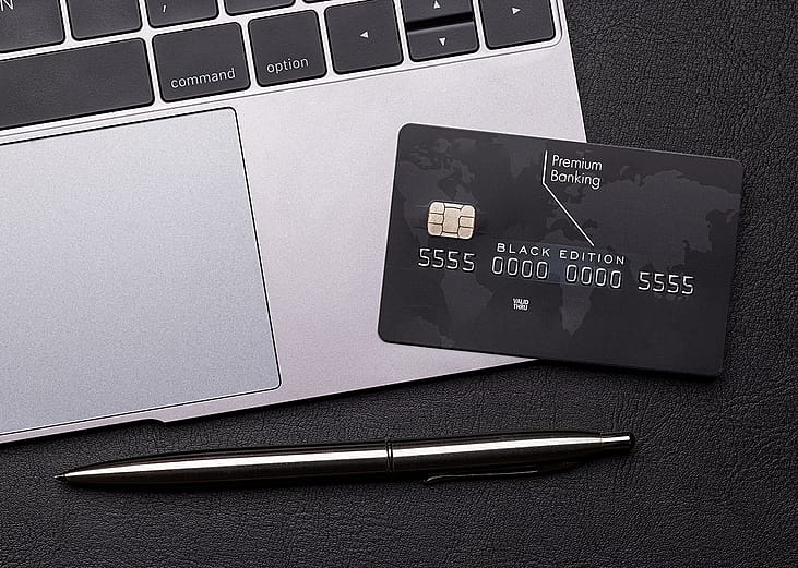 Black premium bank card lays on a silver macbook laptop computer next to the trackpad, with a shiny fancy pen on the table.