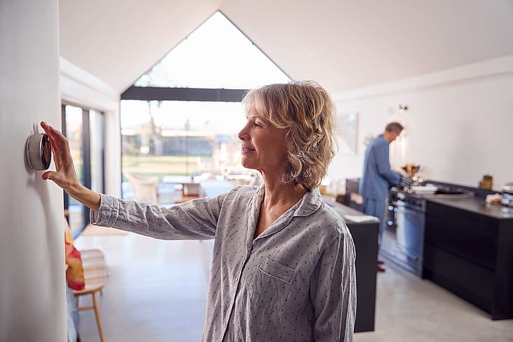 Middle-aged woman wearing pajamas adjusts modern thermostat in a bright house with a vaulted ceiling while a person wearing blue pajamas is seen in the kitchen counter in the background.