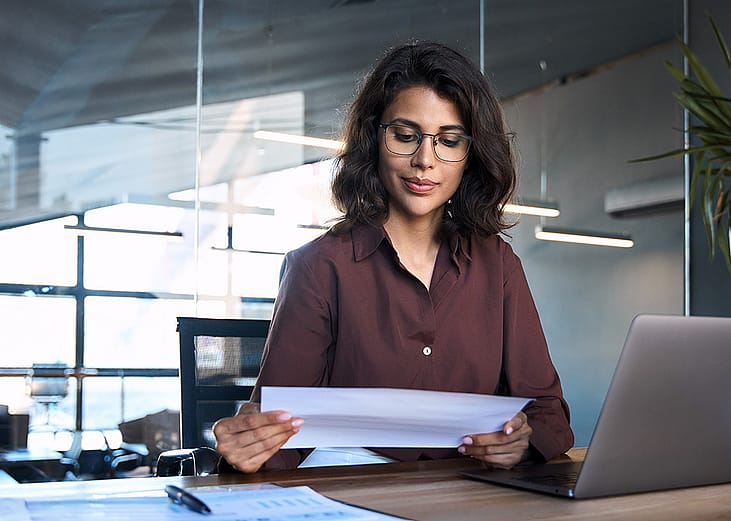 Focused young businesswoman reading financial report document and using a laptop in the workplace.