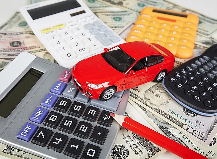 Red toy car surrounded by four calculators and a red pencil sitting on top of U.S. currency to represent the fees required when purchasing a car.
