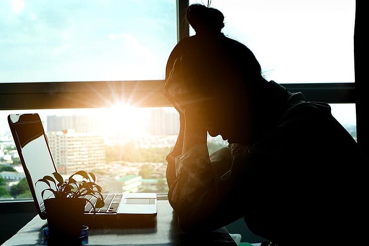 Silhouete of person sitting at desk in front of laptop, with head in hands backlit by sunset shining through office window in background.