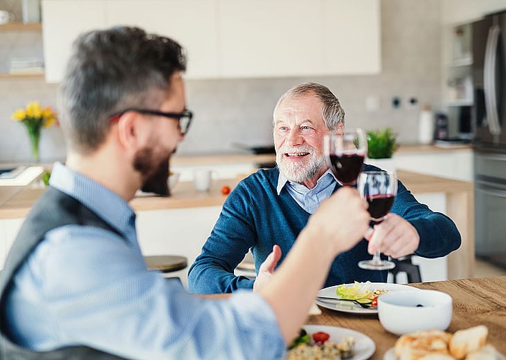 An adult son and his senior father toasting with wine during a light lunch.