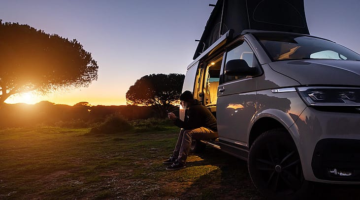 Silhouetted digital nomad sitting in his white transporter camping van, backlit by the setting sun near the California coast.