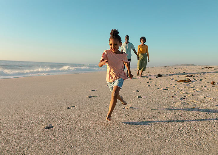 A young girl running on sand against parents walking on a beach.