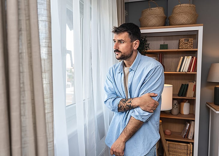 Young sad and depressed anxious looking man wearing a blue button up shirt looking through white curtains out the window.