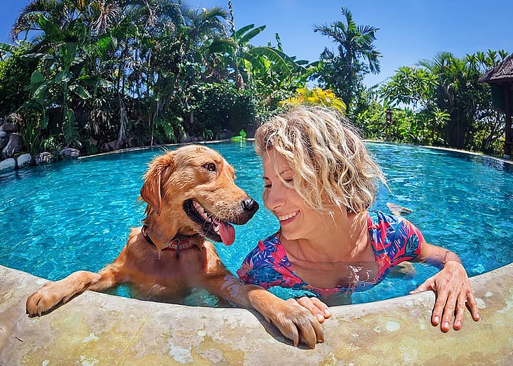 A medium-sized brown dog and blond woman look at each other happily in the pool, with their hands and paws holding onto the edge of the pool.
