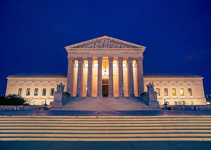 United States Supreme Courts building illuminated at night in Washington DC with steps and plaza in foreground.