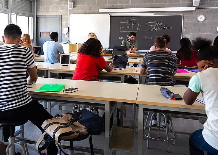 View from back of classroom of multiracial high school students in class using laptops while teacher marks exams.