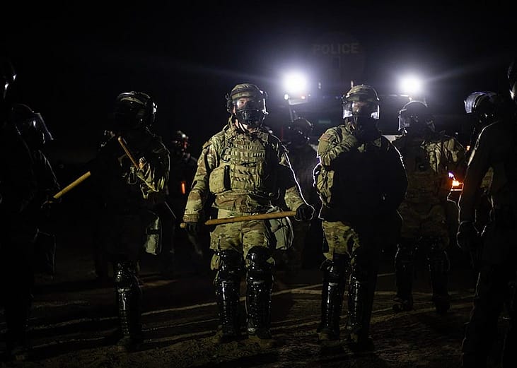 Nighttime view of a line of federal immigration agents in riot gear and face shields facing the camera near Glass House Farms facility in Camarillo, with police spotlights in background.