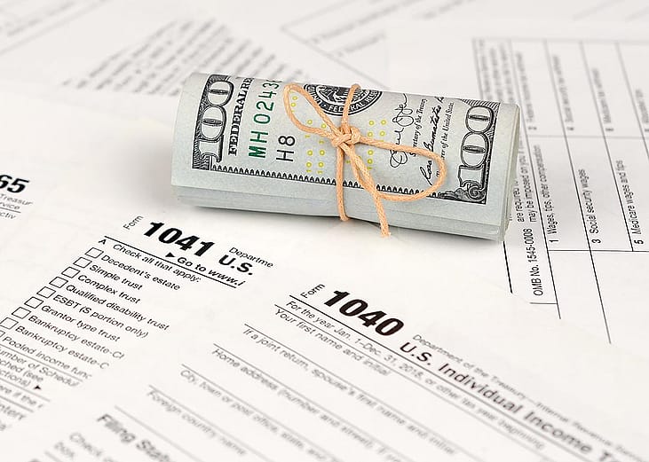 A tied up roll of hundred dollar bills on top of various paper U.S. tax forms like the 1040 and 1041.