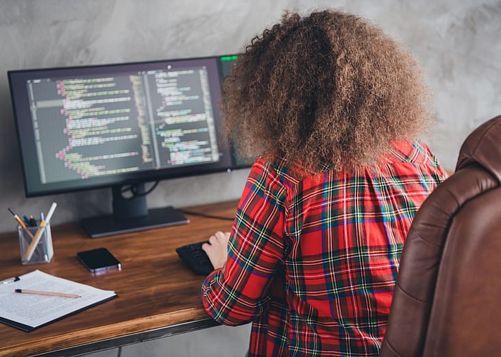 Rear view of a woman with curly hair working at a computer desk displaying code in a casual office setup wearing a checked shirt.