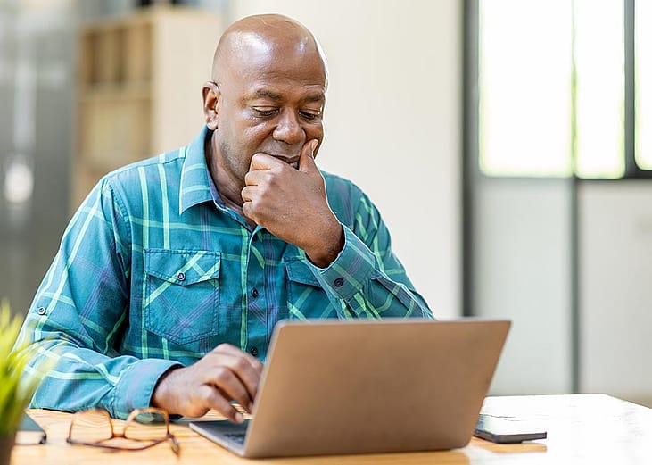 Black bald man wearing teal button up shirt using laptop computer and thinking about question in office.