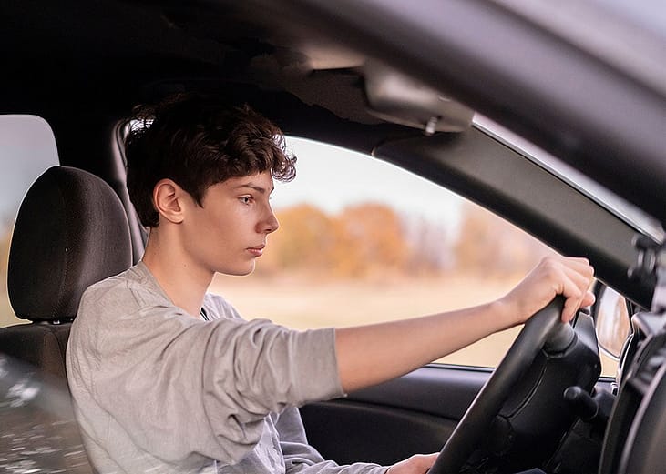 Young male driver holding steering wheel in car seen through the passenger side window.