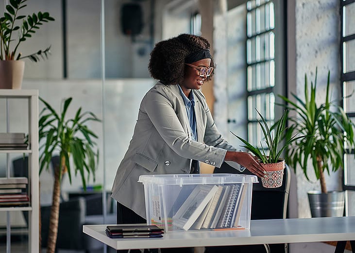 Black woman with glasses placing plants on an office desk with a clear plastic bin full of belongings on table.