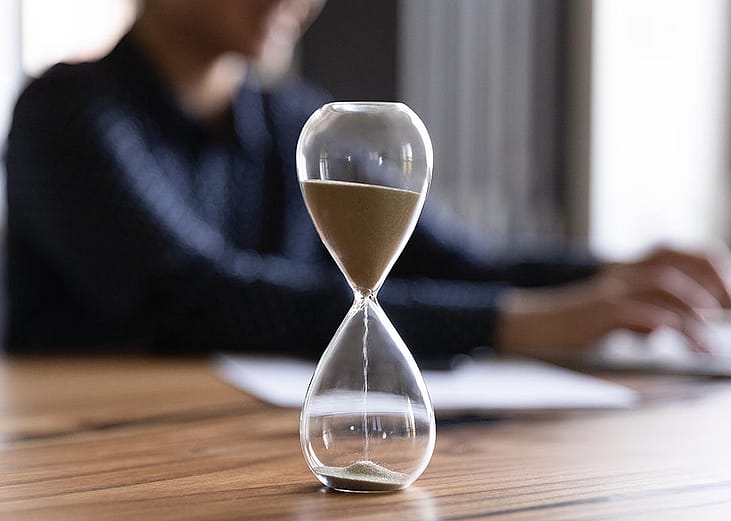 Close up view of hourglass stand on wooden office table measuring time, person working on laptop in the background blurred.