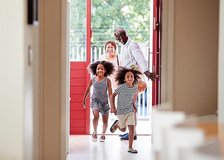 Two cute children with natural hair running into open door of house with grandparents in the background. 