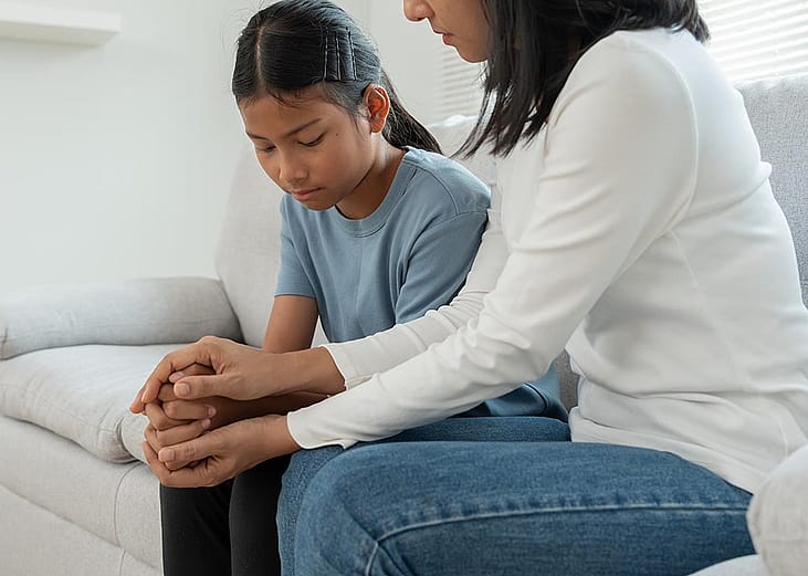 Youth and adult sitting on couch, child has a solemn look and adult is holding their hands.