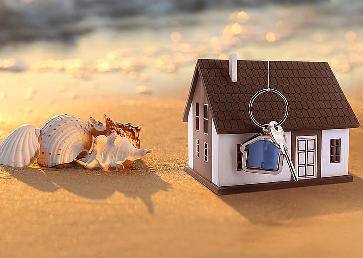 A model of a house with keys on a sandy beach beside seashells.