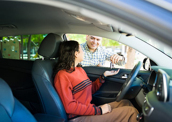 A teenage daughter inside a car receiving its keys from her proud dad.