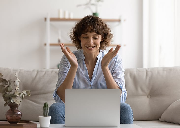 A happy woman sitting on her couch reading good news on her laptop.