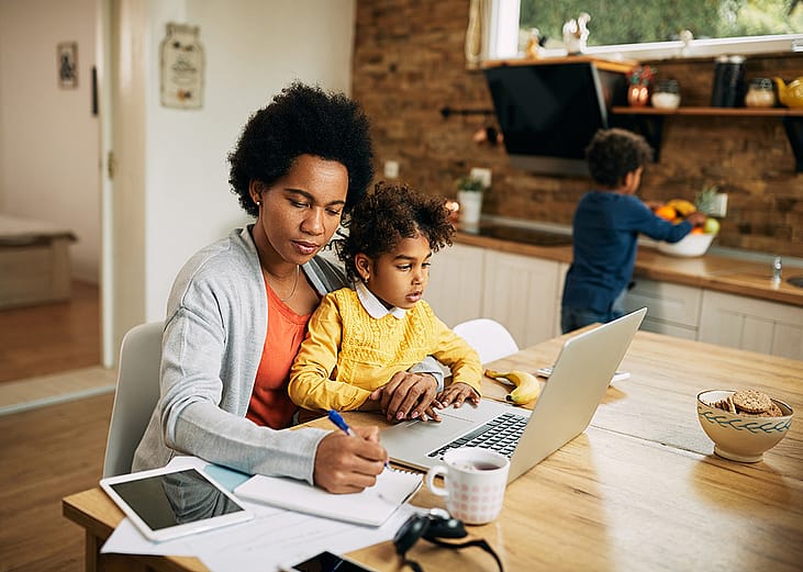 A black mother takes notes as she works at home while her young daughter sits on her lap.