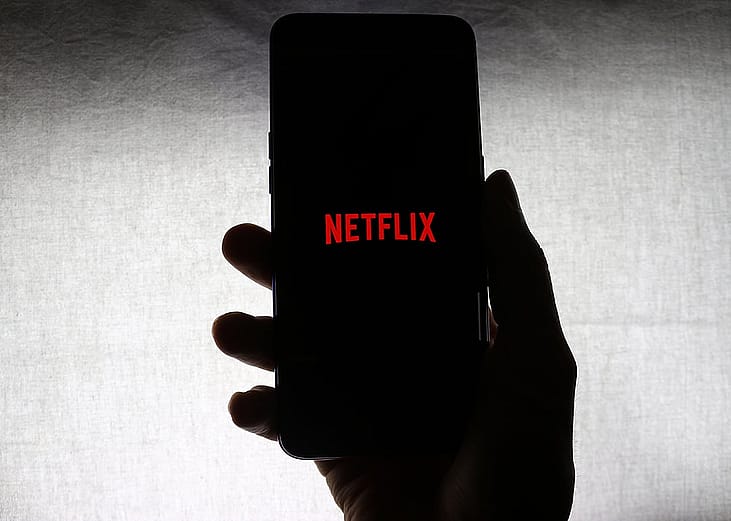 The Netflix logo on the screen of a mobile phone held by a person against a white background.