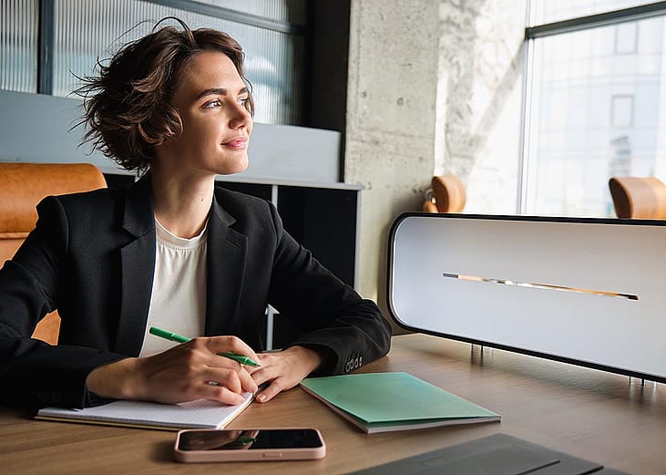 Business woman in office looks toward window thinking while taking notes.