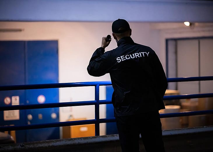 A person standing on a loading ramp outdoors at night wearing dark clothes with the word "SECURITY" displayed on the back of a shirt shines a flashlight toward a white wall with blue doors.