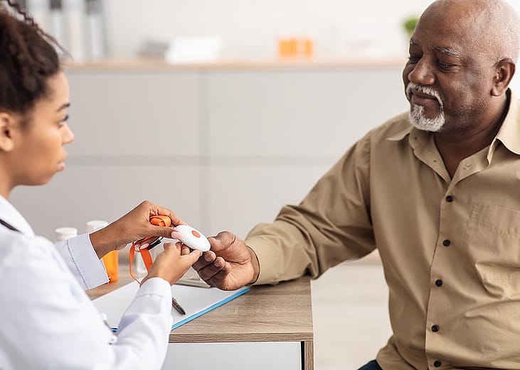 Portrait of black medical worker holding personal alarm button and giving it to mature patient, sitting at table in office, explaining how to use medical alert.