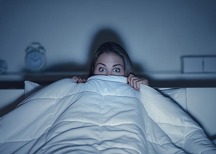 Stylized photo of woman hiding under covers with wide eyes, illuminated by screen from TV.